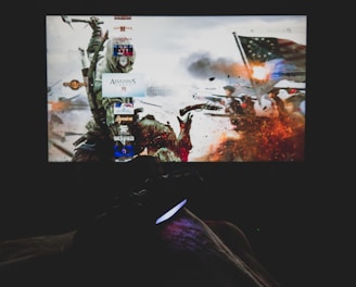 A dimly lit room with a television screen displaying a dynamic, action-packed video game interface featuring a historical battle scene. The foreground shows the silhouette of a gaming console controller held by a person.