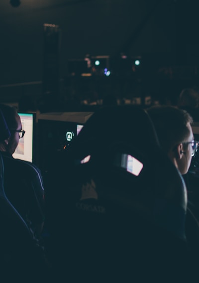 Several individuals are sitting in front of computer screens in a dimly lit room, likely engaging in gaming or working on tasks. The glow of the monitors illuminates their faces, creating a focused and immersive atmosphere. The surroundings suggest a setting like a gaming event or a LAN party.