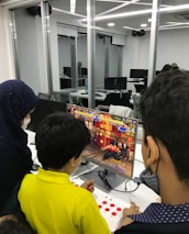 A group of children is gathered around a desktop computer, engaged in playing a colorful, action-packed arcade game displayed on the monitor. The setting appears to be a modern, well-lit office or gaming space with large glass windows, showing multiple computer workstations in the background.