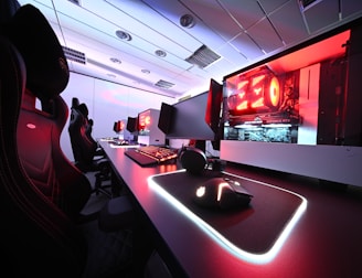 A row of high-end gaming computers on sleek desks, featuring RGB lighting and gaming accessories. The computers are housed in transparent cases with visible components, emitting a red glow. Comfortable gaming chairs with ergonomic design are positioned next to the desks, and the room's ceiling is modern with recessed lighting.