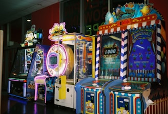 A row of brightly colored arcade game machines is arranged in a line against the wall. Each machine features vibrant lights and designs, with themes ranging from claw games to game show-inspired setups. The area has a playful and inviting atmosphere, suitable for entertainment or amusement centers.