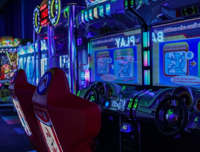 A vibrant arcade environment with several illuminated gaming machines. The image features arcade racing games with visible steering wheels, colorful control panels, and screens displaying game instructions. Bright LED lights in various colors illuminate the machines and surrounding area.