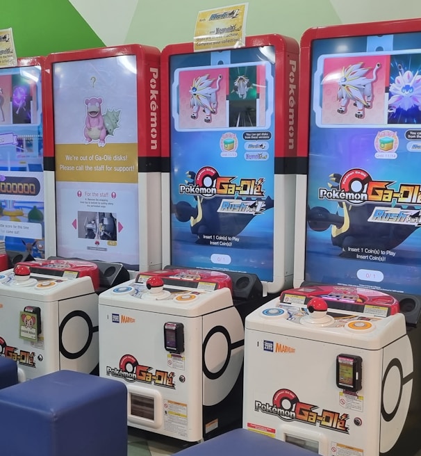 Several arcade gaming machines with multiple screens displaying Pokémon-themed content are positioned in a colorful arcade setting. There are people interacting with the machines; some are seated, and others are standing. The area features different gaming options, including a jumping game with bright, inviting graphics.